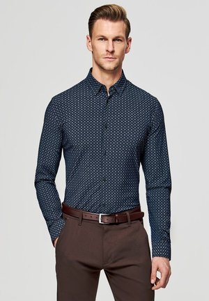 Mężczyzna w dopasowanej granatowej koszuli w wzory zapiętej na guziki, włożonej do brązowych spodni z dopasowanym brązowym paskiem, stojący na jednolitym tle.