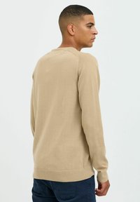 Pull en tricot beige avec un col rond, des poignets et un ourlet à côtes, une texture lisse et des détails sur les panneaux latéraux, porté par une personne de dos.
