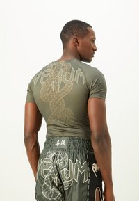 Mand iført en grøn Venum rash guard og matchende shorts med slangedesign, der står sidelæns og viser ryggen og højre profil.