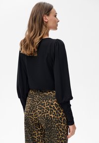 Kobieta z falującymi włosami do ramion, w czarnej bluzce z długim rękawem i spodniami w wysokim stanie w panterkę, stoi na tle białym.