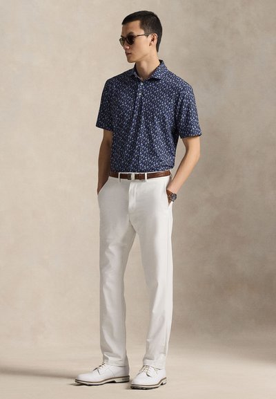 Σκούρο μπλε μπλουζάκι με κοντά μανίκια και φλοράλ μοτίβο, λευκό παντελόνι και λευκά παπούτσια. Αξεσουάρ περιλαμβάνουν γυαλιά ηλίου και καφέ ζώνη.