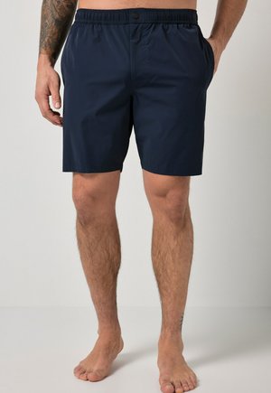 Hombre con pantalones cortos de cintura elástica azul marino, de pie descalzo con las manos en los bolsillos contra un fondo liso.