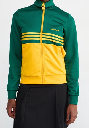 Person iført en grøn og gul Adidas lynlåstrøje med tre horisontale striber og et sort skørt, der står mod en ensfarvet baggrund.