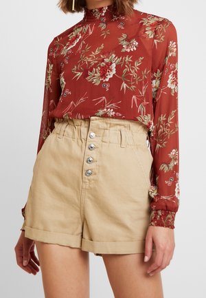 Kvinde bærer beige højtaljede shorts med fire sølvknapper og en rød blomster-print langærmet bluse med flæsekrave og manchetter.