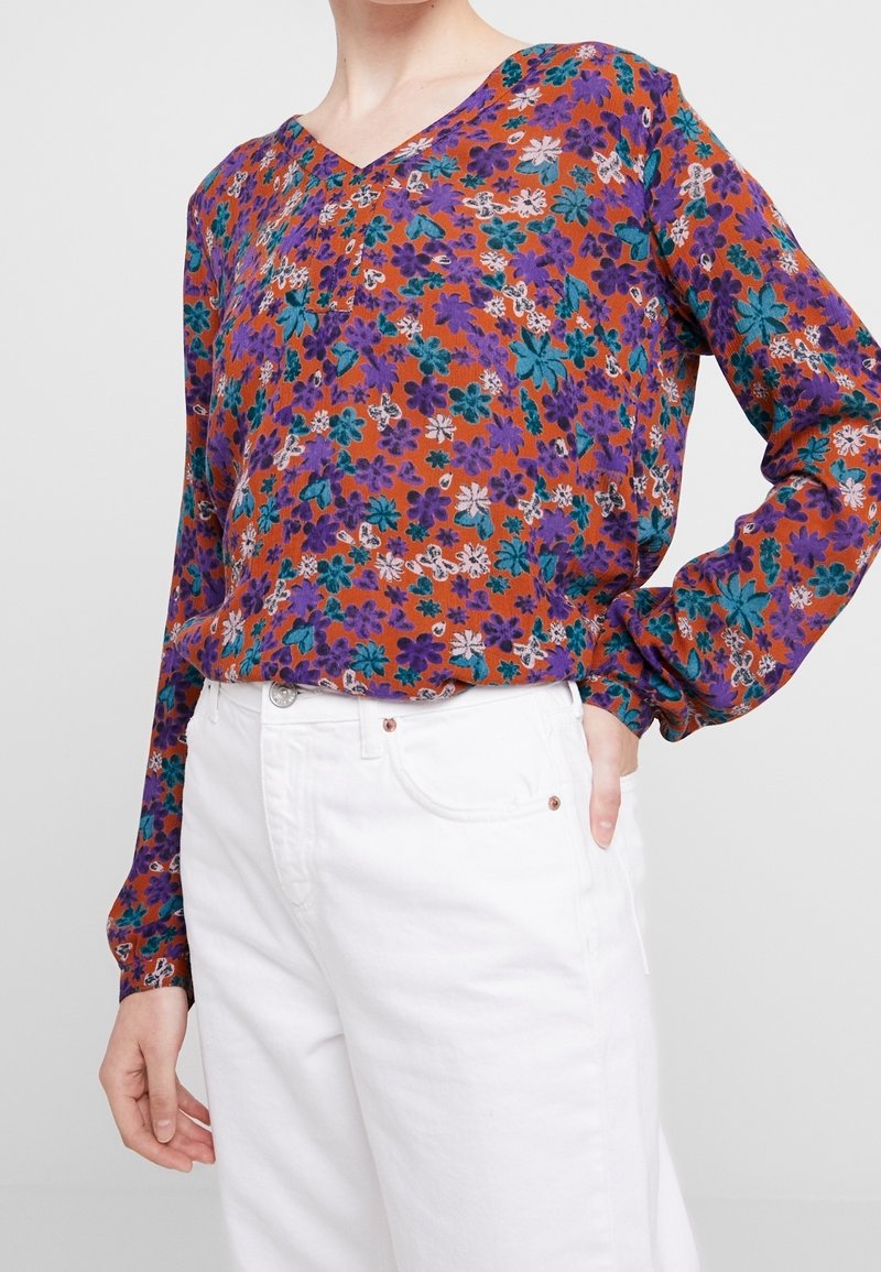 Blusa floreale multicolore con scollatura a V, caratterizzata da tonalità di arancione, viola e verde acqua. Maniche lunghe con polsini e tessuto leggero.