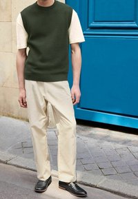 Personne portant un gilet en tricot vert olive sur un t-shirt crème, un pantalon crème et des mocassins noirs, debout près d'une porte bleue dans une rue pavée.