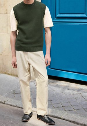 Personne portant un gilet en tricot vert olive sur un t-shirt crème, un pantalon crème et des mocassins noirs, debout près d'une porte bleue dans une rue pavée.