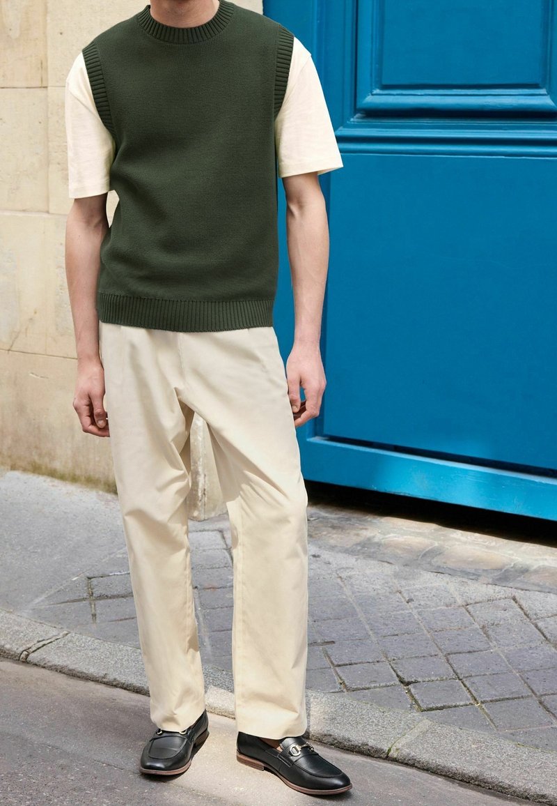 Personne portant un gilet en tricot vert olive sur un t-shirt crème, un pantalon crème et des mocassins noirs, debout près d'une porte bleue dans une rue pavée.