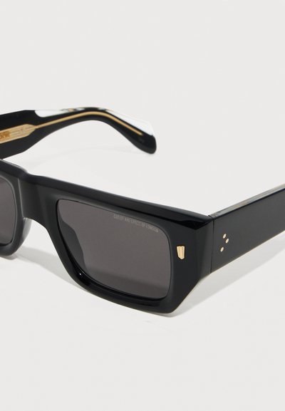 Zwarte rechthoekige zonnebril met donkere lenzen, gouden accenten op de pootjes en een klein gouden embleem vlak bij het scharnier op een witte achtergrond.