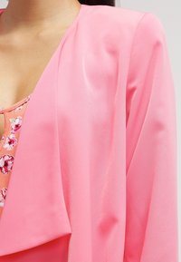 Růžový šifonový kardigan s volným střihem a měkkou texturou, pod nímž se nachází top s květinovým vzorem.