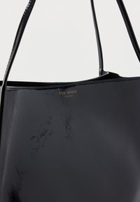 Czarna torebka typu tote z lakierowanej skóry, z dwoma uchwytami, o fakturowanej powierzchni i złotym logo "Ted Baker London" na przodzie.