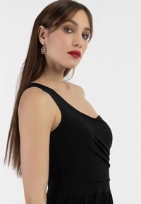 Rochie neagră fără mâneci, cu detaliu drapat în față și textură netedă. Afișată cu cercei aurii din fir și machiaj minimal.