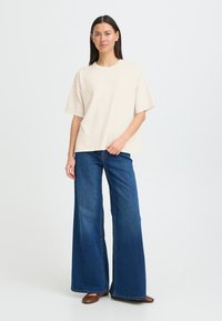 T-shirt beige oversize à manches courtes associé à un jean bleu en denim à jambes larges. Texture lisse, coupe décontractée et légère nervure sur le t-shirt.