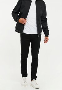 Veste bomber noire avec fermeture éclair, poignets côtelés et rayures, associée à un jean noir ajusté et des baskets blanches. Design simple et moderne.