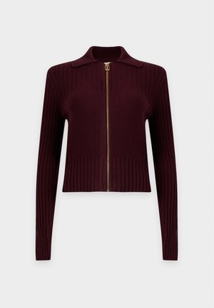 Donna in piedi con le mani dietro la schiena, indossa un maglione bordeaux con zip, jeans blu e tacchi neri aperti davanti su sfondo bianco semplice.