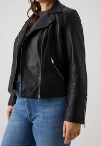 Giacca da moto in pelle nera con zip argentate, colletto a scatto e dettagli a coste sui polsini. Indossata sopra una maglietta grigia e jeans blu.