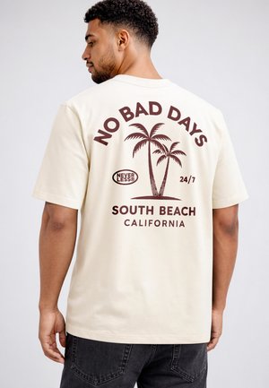 Man facing away wearing cream t-shirt with brown text and palm tree graphic saying "No Bad Days South Beach California"