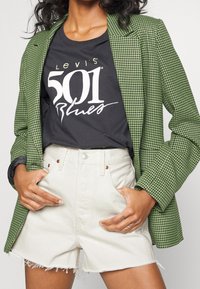 Person som bär en grön hundtandsmönstrad kavaj, svart Levi's 501 Blues t-shirt och högmidjade vita denimshorts med fransade kanter.