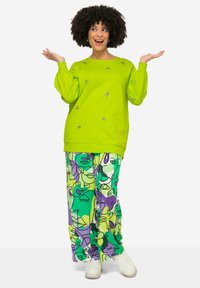 Sudadera verde de gran tamaño con mangas abullonadas y detalles plateados, combinada con pantalones de pierna ancha estampados que presentan rostros abstractos en verde y púrpura.