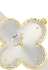 Montre dorée avec un cadran en argent en forme de fleur, dotée de aiguilles dorées et d'un logo. Le bracelet blanc lisse complète le design.