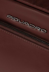 Accesorio de cuero burdeos con cremallera metálica y logo plateado "Piquadro", destacando su textura suave y diseño refinado.