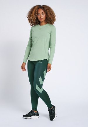 Vrouw met krullend haar die een lichtgroene top met lange mouwen, donkergroene legging met chevronpatroon en zwarte sportschoenen draagt, staat ontspannen.