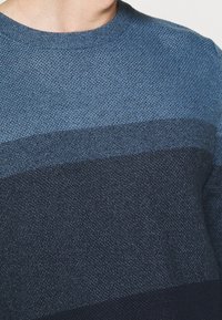 Blåstickad tröja med en graderad design och texturerad yta, som övergår från ljusblått till mörk marinblått i botten.