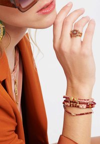 Bague en or avec une pierre rouge, plusieurs bracelets empilés dans des teintes de rose et de rouge, et de grandes boucles d'oreilles vertes ; mannequin portant un blazer de couleur rouille.