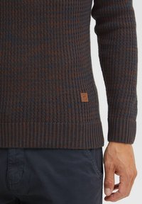 Knit sweater with a textured pattern in dark blue and brown tones. Features a ribbed hem and a small leather logo tag on the side.