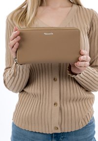 Nainen, joka pitää käsissään beigeä ribbineulosta neuletakkia ja ruskeaa vetoketjullista lompakkoa, jossa näkyy tuotemerkki "Tamaris".