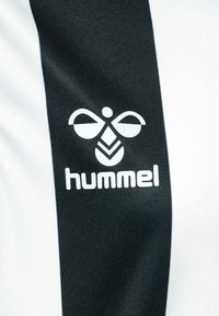 Svart och vit idrottströja med Hummel-logotypen. Tyget verkar slätt, med en glansig yta och djärva vertikala ränder.