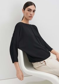Blusa nera, ampia, con scollo tondo, maniche tre quarti e dettaglio centrale cucito, indossata sopra pantaloni chiari.