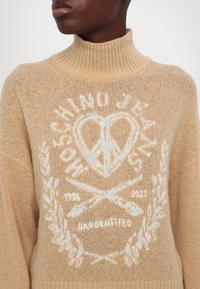 Pull à col roulé en tricot beige avec un motif blanc représentant un symbole de paix en forme de cœur, des aiguilles à tricoter croisées, et les textes "Chino Jeans" et "Fait main".