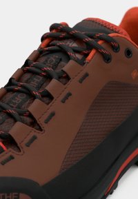 Καφέ μποτάκι πεζοπορίας με μαύρες λεπτομέρειες, υφή στην επιφάνεια, κόκκινες κορδόνια και ανθεκτική λαστιχένια σόλα. Διαθέτει ενισχυμένη μύτη και ραφές σε ζιγκ-ζαγκ.