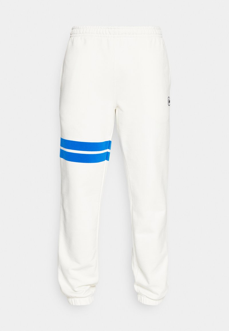 Pantaloni della tuta bianchi in morbido tessuto, con strisce blu e un logo circolare sulla coscia. Vita e polsini elasticizzati per una vestibilità affusolata.