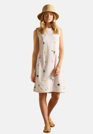 Baumwollkleid mit Bestickung von Blumen, Palmen und Luftballons, mit einem welligen Saum und ärmellosem Design. Getragen mit gelben Sandalen.