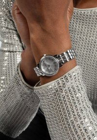 Silberner Chronograph mit rundem Ziffernblatt, metallischem Armband und Kristallakzenten, getragen an einem Handgelenk neben einem strukturierten silbernen Kleidungsstück.