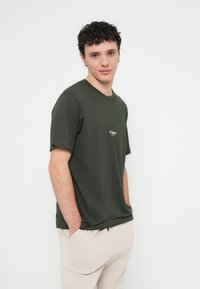 Dunkelgrünes kurzärmliges T-Shirt aus Baumwolle mit kleinem weißem Logo auf der Brust. Das Modell steht mit den Händen in den Taschen und trägt beige Hosen.