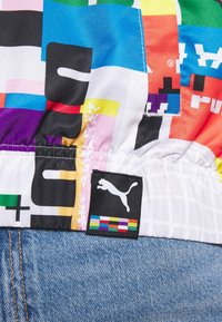 Kolorowa tkanina w wzory z mieszanką geometrycznych kształtów. Zawiera czarną metkę z logo Puma z białą sylwetką i kolorowymi akcentami.