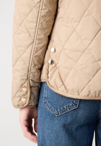 Beige kviltet jakke med guld trykknapper på ærmekant og side, parret med blå denim jeans, der viser baglommen.
