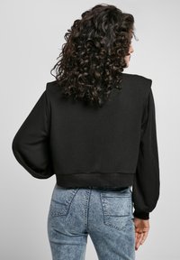 Femme aux longs cheveux bouclés, portant un sweat-shirt noir court et un jean délavé bleu clair, de dos contre un fond uni.