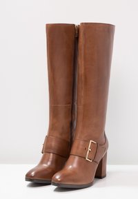 Botas de cuero marrón hasta la rodilla con una textura suave, cremallera lateral y un acento de hebilla cuadrada en el tobillo. Diseño de tacón cuadrado.