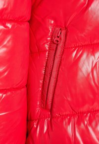 Röd puffjacka med en glansig yta, kviltade sektioner och en dragkedjeficka på sidan. Slät textur med rundade kanter.