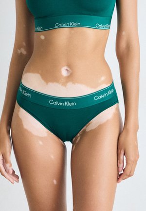 Groene Calvin Klein bralette en bijpassende bikini, gemaakt van zachte stof, met een logoband en een gladde textuur in verschillende huidskleuren.