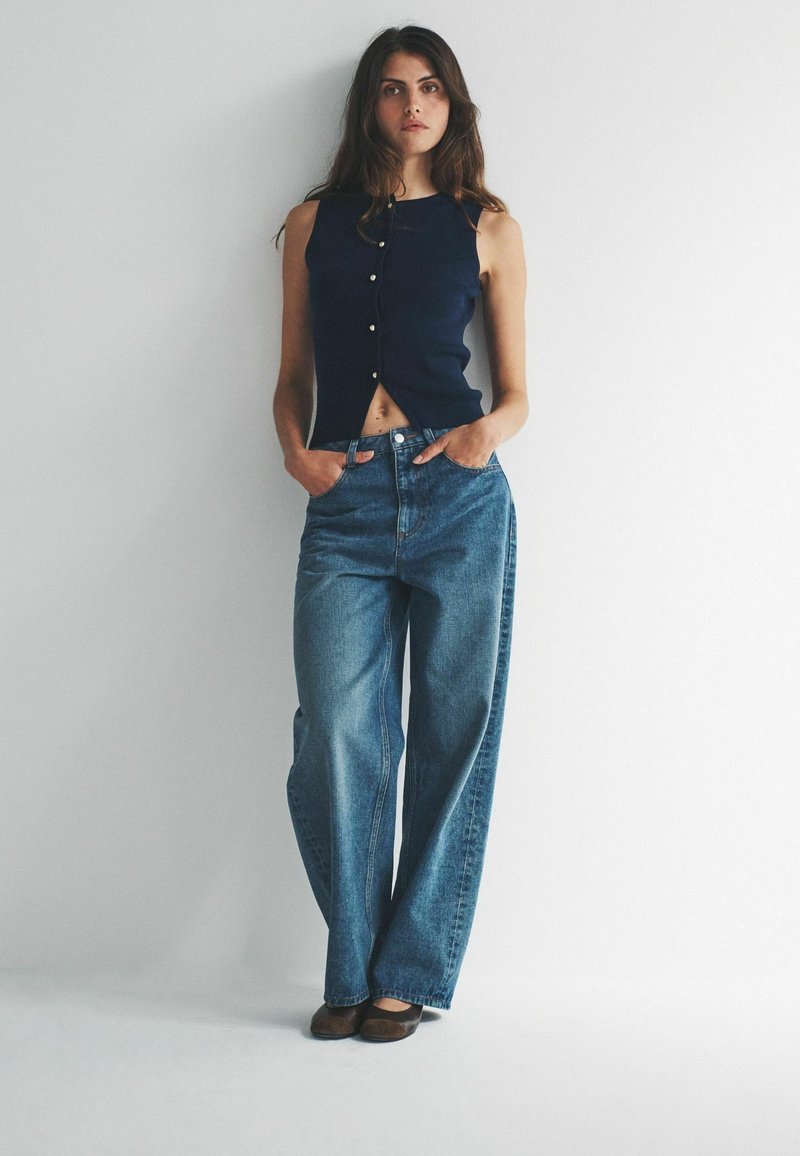 Tmavomodrý top bez rukávov s gombíkmi, kombinovaný so svetlomodrými širokými džínsami. Model stojí s rukami v vreckách na bielom pozadí.