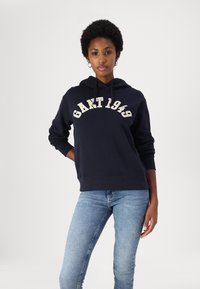 Haut à capuche bleu marine en coton, doté d'une poche kangourou à l'avant, d'une capuche avec cordon de serrage et de la mention "GANT 1949" en tissu crème.