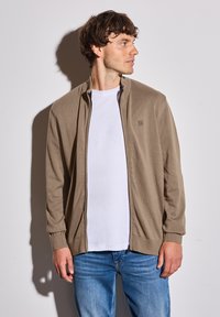 Světle hnědý kardigan na zip s vroubkovanými manžetami a límcem, nošený přes bílý tričko. Tkanina vypadá měkce a lehce.