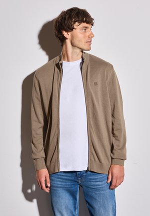 Cardigan zip-up marrone chiaro con polsini e colletto a coste, indossato sopra una t-shirt bianca. Il tessuto appare morbido e leggero.