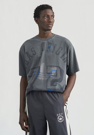 Hall hallikate graafilise trükiga suur T-särk, millel on sinised aktsendid ja tekst "72". Ühildatud tumedate hallide pükstega, millel on valged triibud ja logo.