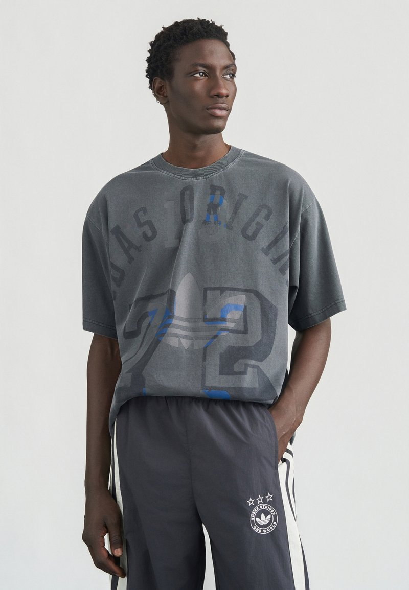 Γκρι oversized T-shirt με γραφικό σχέδιο, που περιλαμβάνει μπλε αποχρώσεις και το κείμενο «72». Συνδυάζεται με σκούρο γκρι παντελόνι με λευκές ρίγες και λογότυπο.